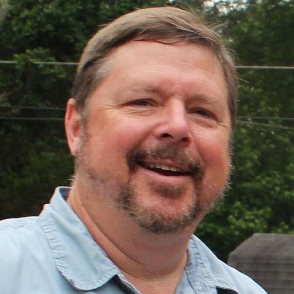 A white-presenting male with light brown hair and a light beard, smiling, wearing a light blue button-down shirt.