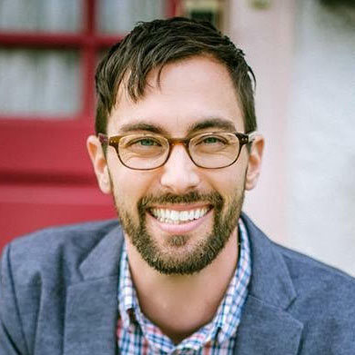 A white-presenting male with brown hair, a beard and brown glasses, smiling, wearing a jean colored suit jacket and blue and red button-down shirt.