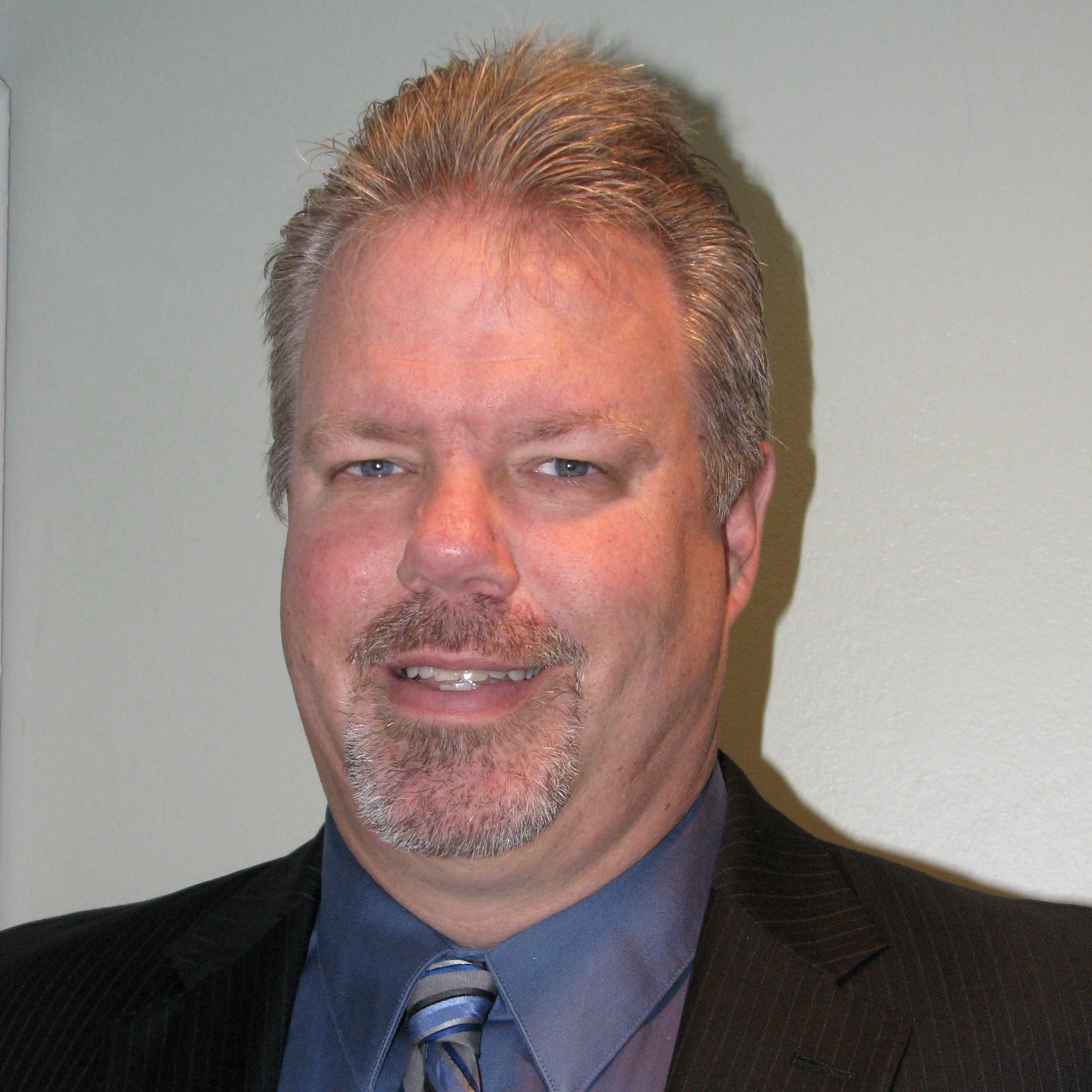 A white-presenting male with salt and pepper hair, a mustache and goatee, with blue eyes, wearing a black suit jacket and blue button-down shirt.